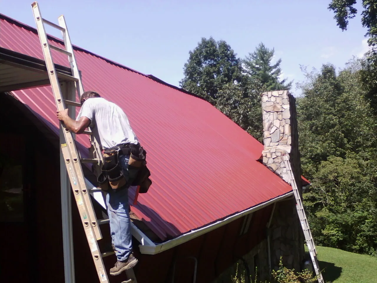 Roofer on a ladder beside a red metal roof and stone chimney — Soffit & Fascia Repair by Roofing Brothers in Elm Grove